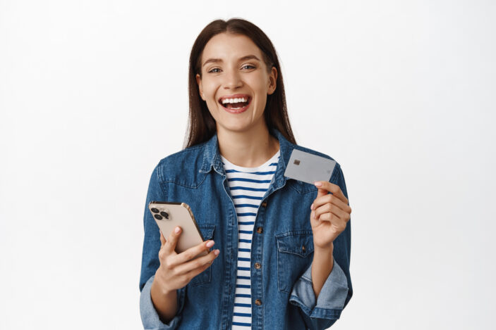 Happy woman holding smartphone, showing credit card, smiling satisfied and laughing, advertising online shopping sale, buying smth on mobile phone, white background